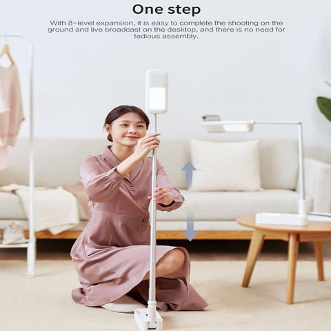 Woman in pink dress adjusting height of 3-in-1 foldable LED fill light smartphone stand in cozy living room