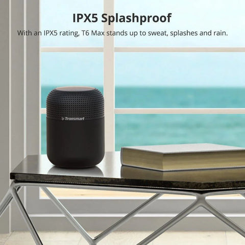 Black Tronsmart T6 Max Bluetooth speaker on a table with a book, by a window showing ocean view