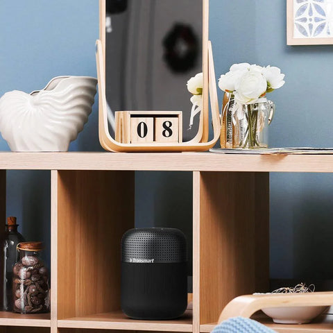 Black Tronsmart Bluetooth speaker on wooden shelf with decorative white vase and flowers