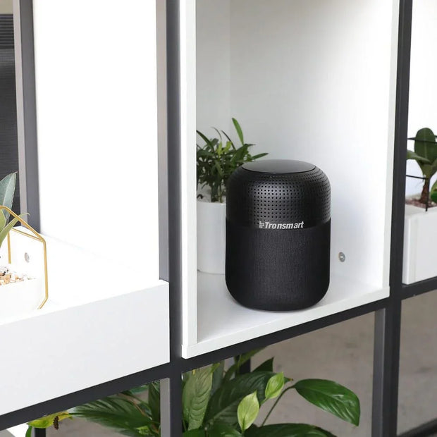 Black Tronsmart Bluetooth speaker placed on a white shelf with green plants in the background
