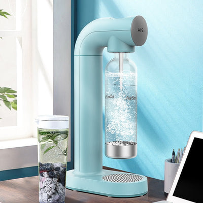 Light blue AirS soda machine carbonating water in a clear bottle on kitchen counter beside a glass of infused fruit water