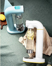 Two modern carbonated water makers with clear bottles, one with lemon and cucumber slices, on green textured surface