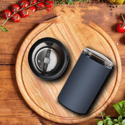 Black electric coffee grinder disassembled on round wooden cutting board with fresh cherry tomatoes and herbs