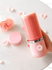 Hand holding pink electric portable blender with fresh pink smoothie on marble table with rose petals