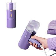 Purple foldable handheld garment steamer with transparent water tank, held by hand, next to folded purple clothes