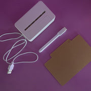 White USB cable, white electronic device, white pen, and brown card on purple background
