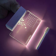 Light message writing board with calendar and floral design illuminated in purple, pen beside it