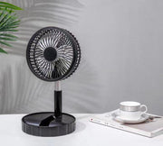 black portable telescopic folding fan on white table next to book and cup with plant shadow on gray wall